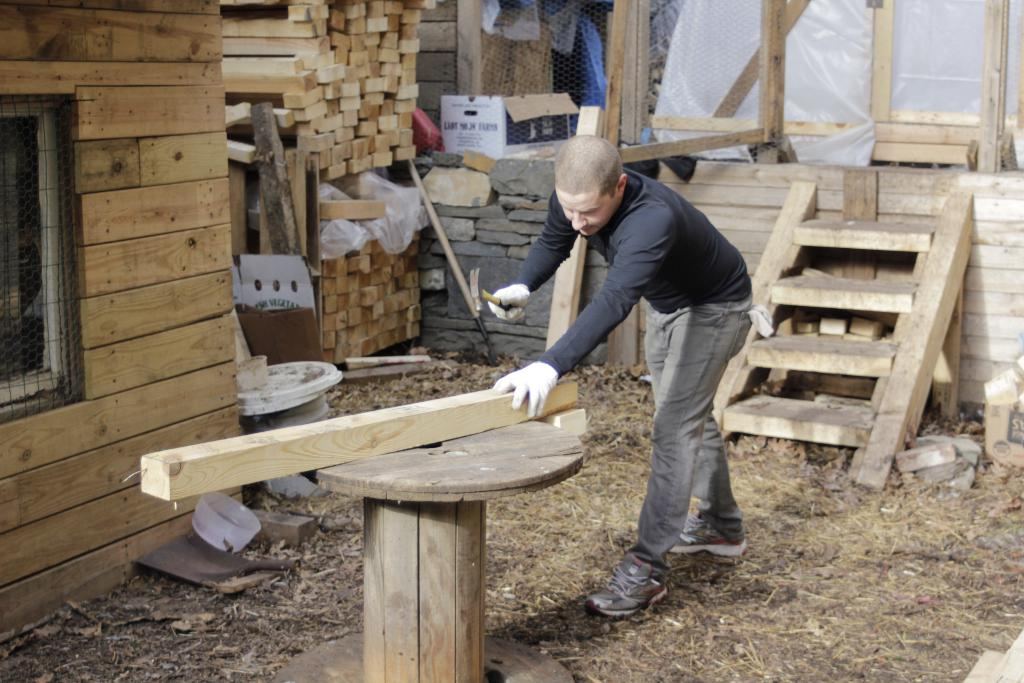A builder measuring timber beside a workshop full of stacked boards