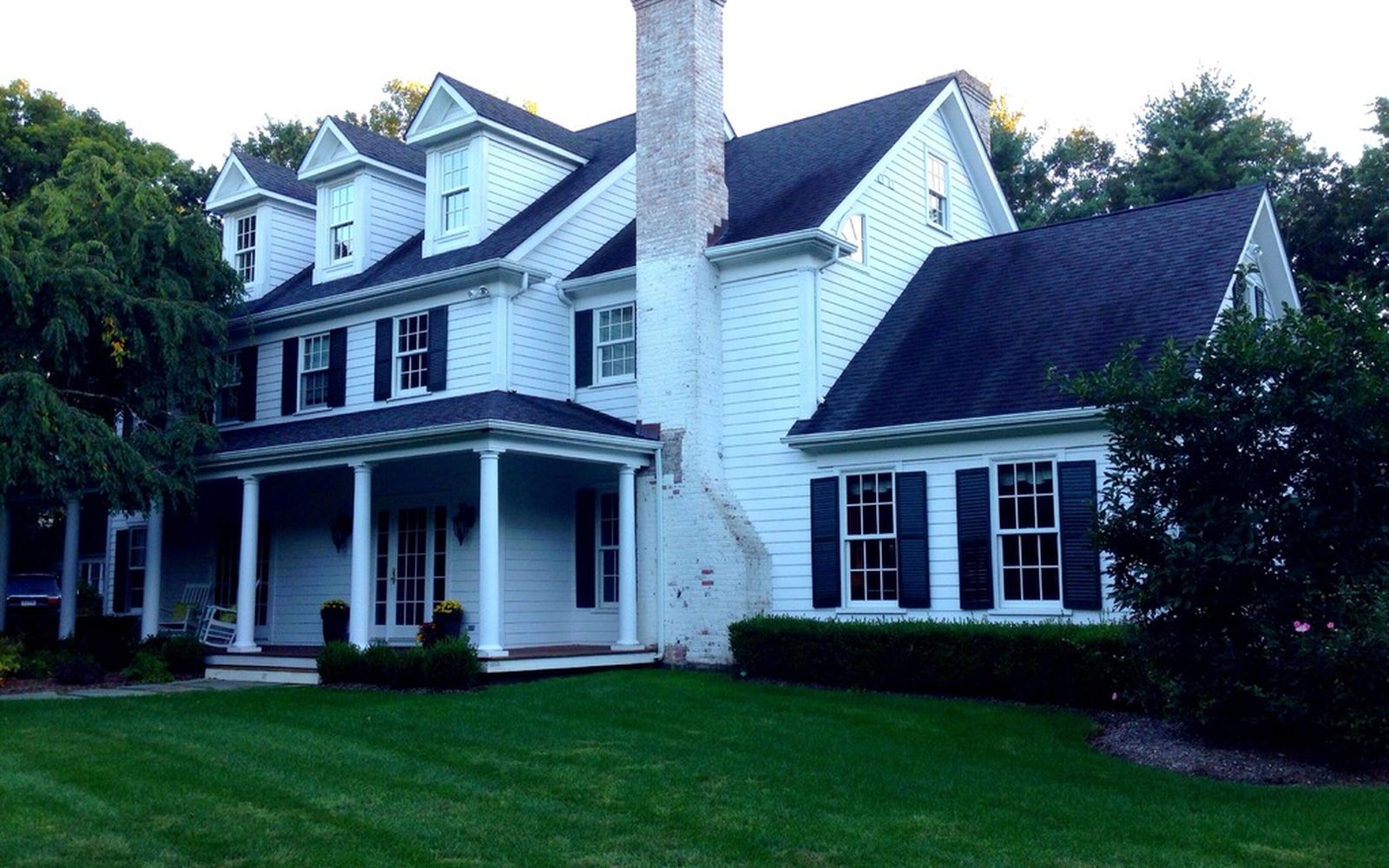 A white colonial-style family home with dark shutters, a wide porch and a green lawn
